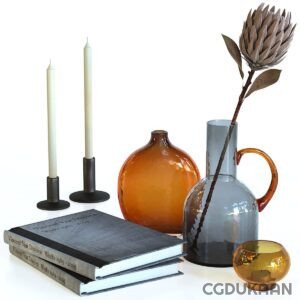 A still life composition with two cream-colored candles on black holders, a stack of three books titled "Vincent Van Duysen Works 1989-2009", an orange round vase, a grey pitcher with brown handle holding a dried protea flower, and a small amber glass sphere.